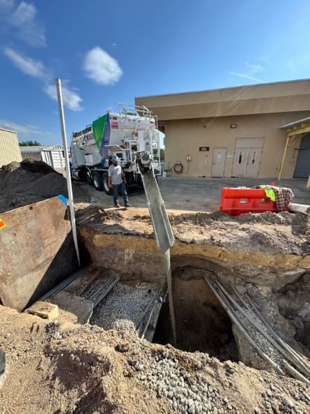 Quality-Crete volumetric mixer placing concrete through a chute into an excavated utility trench while a crew member watches