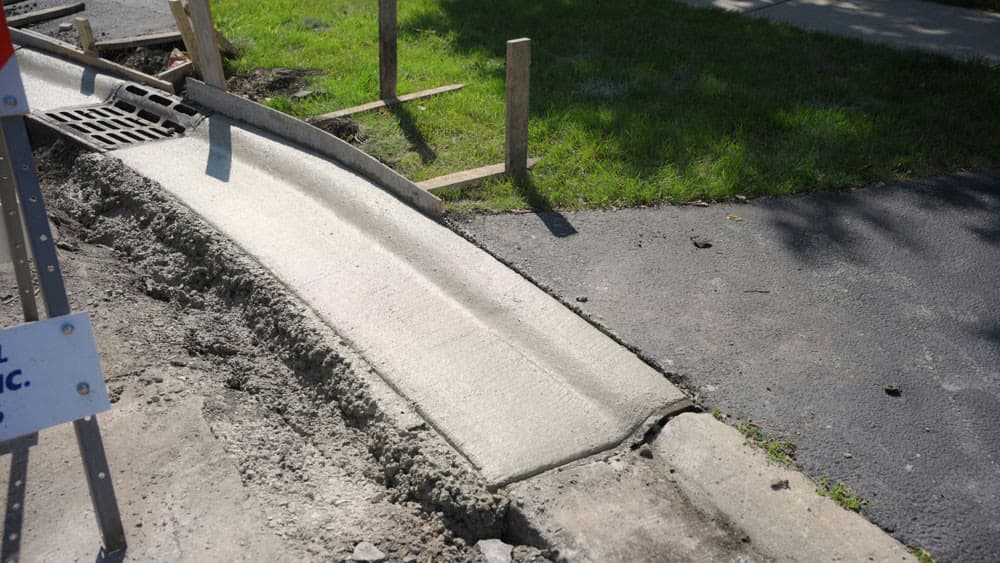 Freshly poured concrete curb cut and driveway apron beside a storm drain grate, backfilled with loose soil