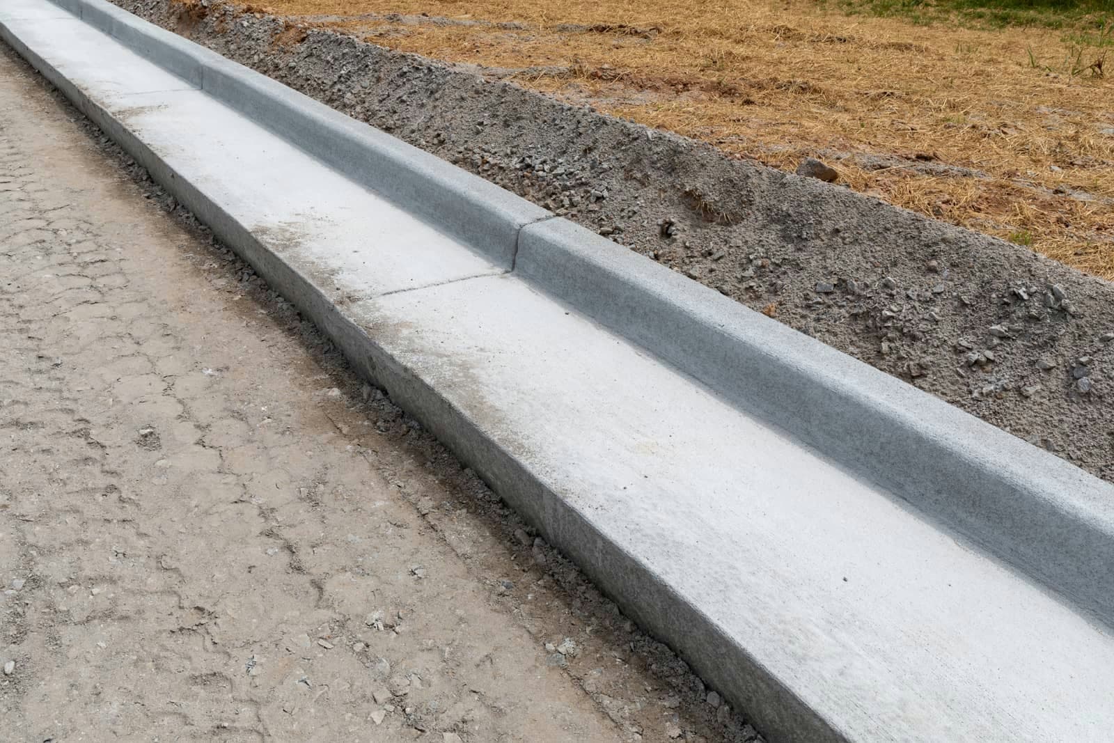 Concrete curb and gutter along an unpaved road, backfilled against a grassy field