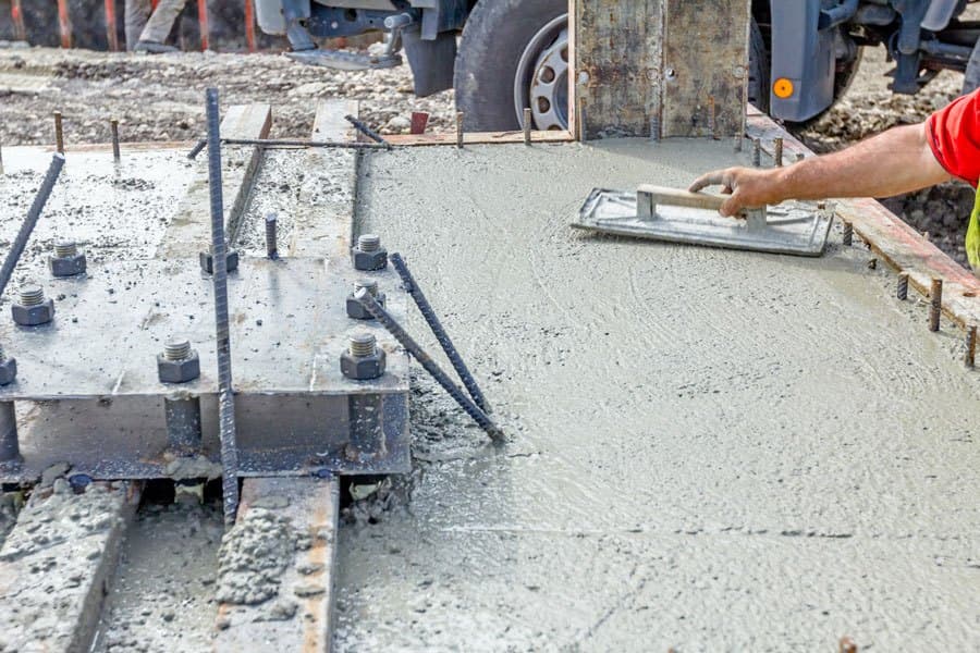 Worker trowel-finishing fresh grout around anchor bolts on an equipment foundation, with a mixer truck alongside
